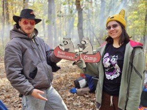 Two people (Sarge and Gator) posing with a Bear Essentials sign. The sign features two raccoons rowing a canoe.